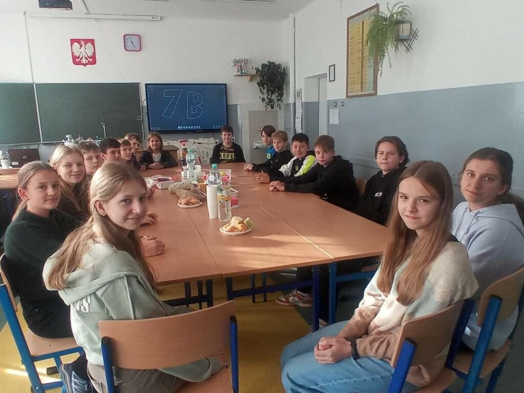 Grupa uczniów siedzących dookoła zsuniętych stolików, na których znajdują się przekąski