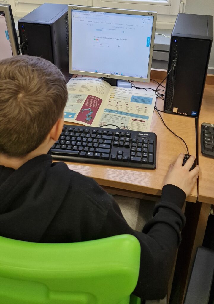 Uczeń w czarnej bluzie pracuje przy komputerze stacjonarnym, na ekranie widnieje zadanie z wyborem pasujących emoji, a przed monitorem leży otwarty podręcznik do informatyki.