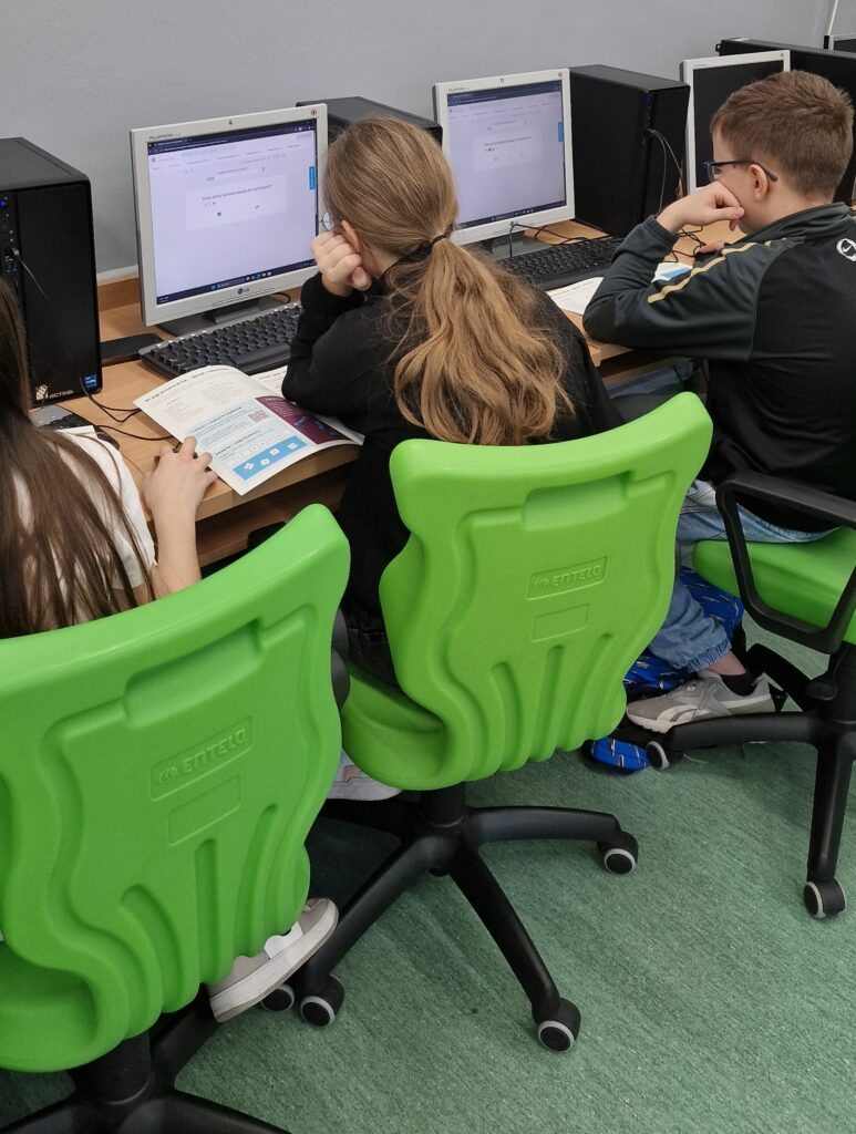 Troje uczniów siedzi przy stanowiskach komputerowych w pracowni, na monitorach widać zadania z emoji, a dzieci pracują z otwartymi podręcznikami, siedząc na charakterystycznych zielonych krzesłach.