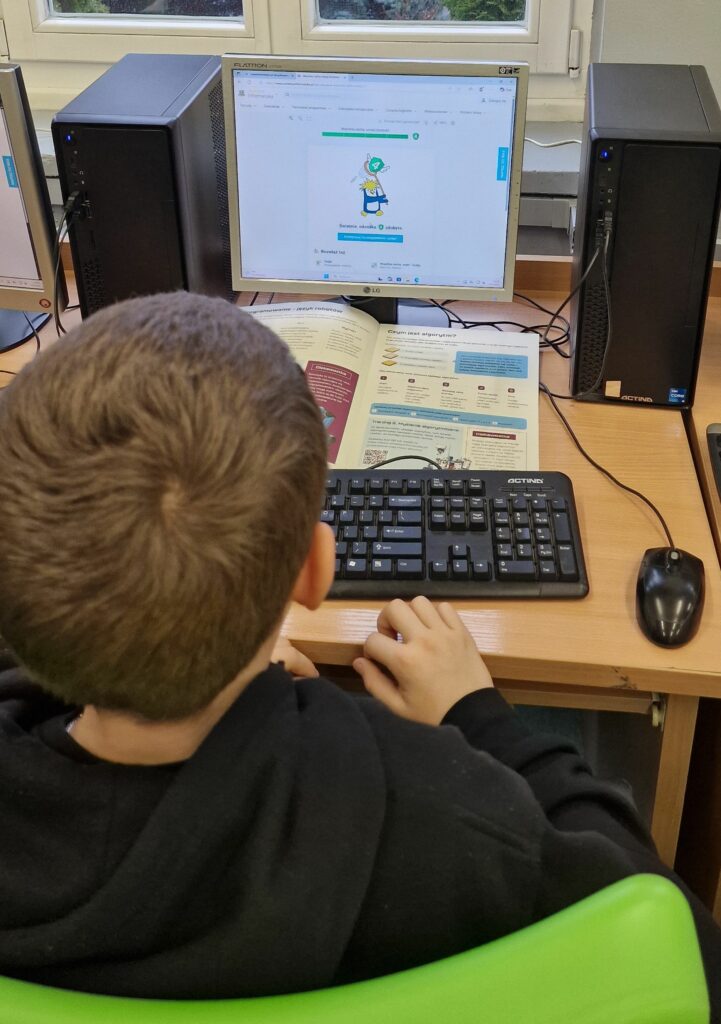 Uczeń siedzi przy szkolnym komputerze, pracuje z otwartym podręcznikiem informatyki i wykonywanym na monitorze zadaniem, na ekranie widać prostą grafikę oraz pasek postępu nauki.