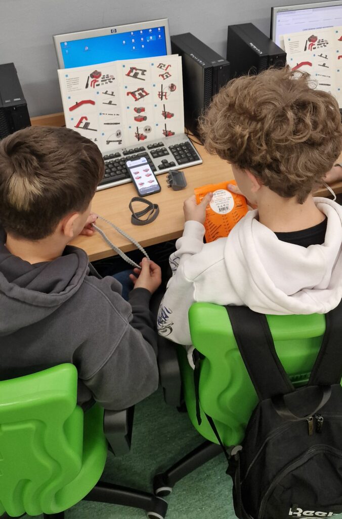Dwóch uczniów siedzi tyłem przy biurku komputerowym, przed nimi stoi monitor z niebieskim ekranem pulpitu, a na biurku rozłożona jest obrazkowa instrukcja konstrukcji robota oraz smartfon z aplikacją pokazującą części.