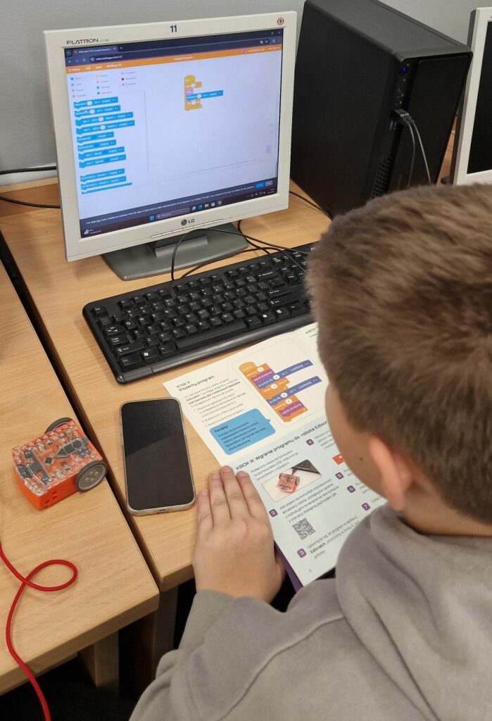 Uczeń przy biurku komputerowym programuje w środowisku blokowym na monitorze, obok klawiatury leży smartfon i mały pomarańczowy robot edukacyjny.