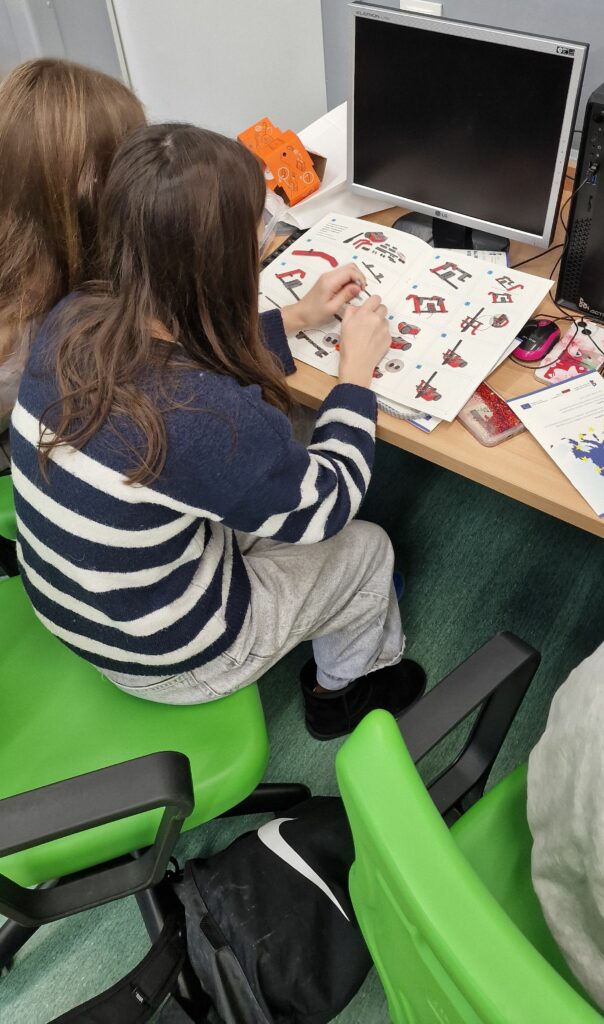 Dwie uczennice siedzą obok siebie na zielonych krzesłach, na biurku przed nimi leży otwarta instrukcja z rysunkami czerwono–szarego robota, jedna z dziewcząt trzyma w dłoniach mały element konstrukcyjny.