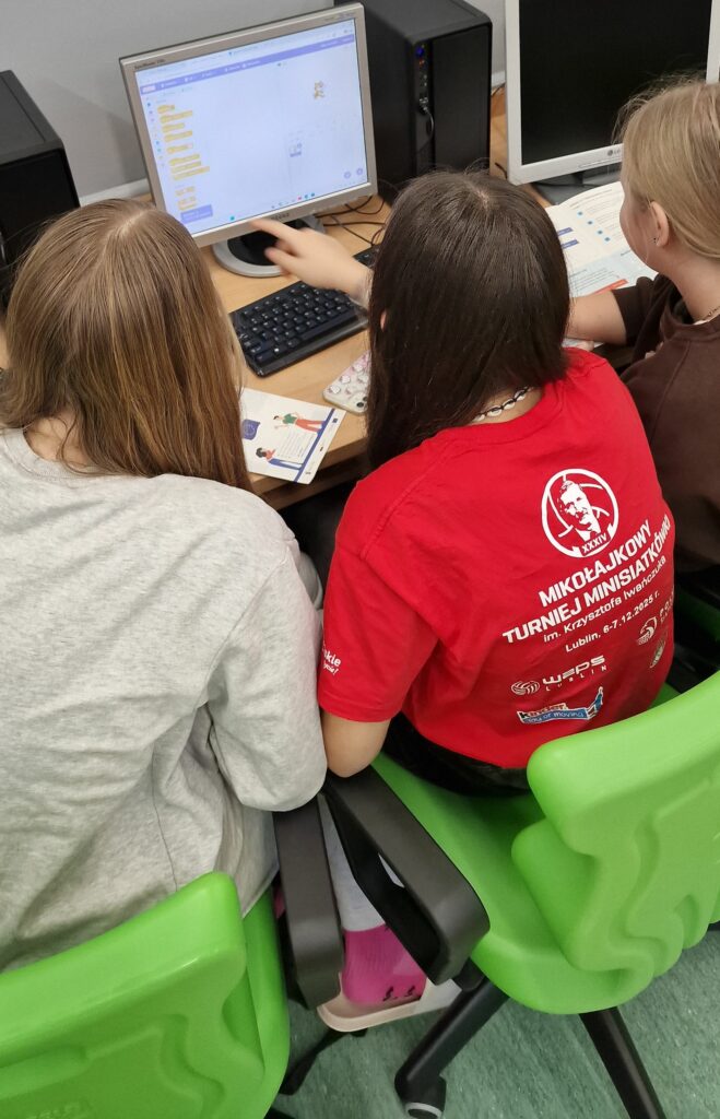 Trzy uczennice siedzą przy jednym stanowisku komputerowym na zielonych krzesłach, wspólnie patrzą na monitor z otwartym środowiskiem Scratch i dyskutują nad kodem.