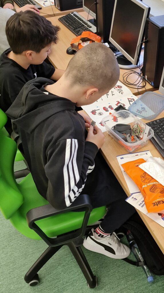 Dwaj uczniowie siedzą obok siebie przy biurku komputerowym, jeden z nich składa elementy robota z zestawu części w plastikowym pudełku, na stole leży rozłożona obrazkowa instrukcja.