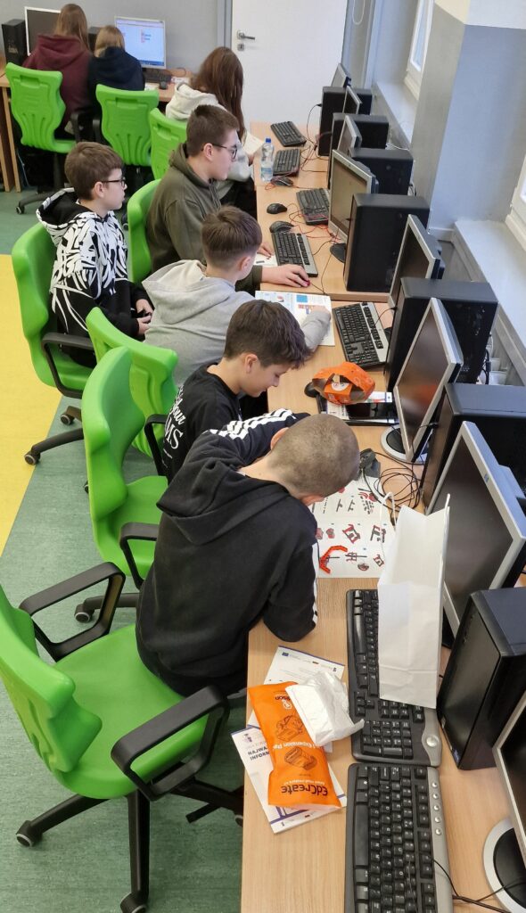 Kilku uczniów siedzi w rzędzie przy stanowiskach komputerowych w pracowni informatycznej, część z nich pracuje przy klawiaturach, inni pochylają się nad instrukcją i elementami robota, na krzesłach znajdują się zielone plastikowe oparcia.