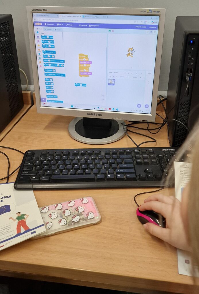Uczeń obsługuje komputer z otwartym środowiskiem Scratch, na ekranie widoczne są bloczki skryptu i kot–duszek, na biurku leżą smartfon w etui z motywem Hello Kitty i materiały edukacyjne.