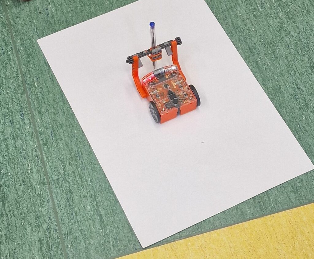 Mały pomarańczowy robot edukacyjny na dwóch kołach stoi na środku białej kartki papieru położonej na zielonej podłodze, na robocie zamocowany jest pionowy uchwyt z długopisem.