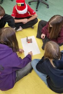 Grupa pięciorga uczniów siedzi po turecku na podłodze wokół dużej kartki papieru, na której znajduje się pomarańczowy robot rysujący po wyznaczonych liniach.