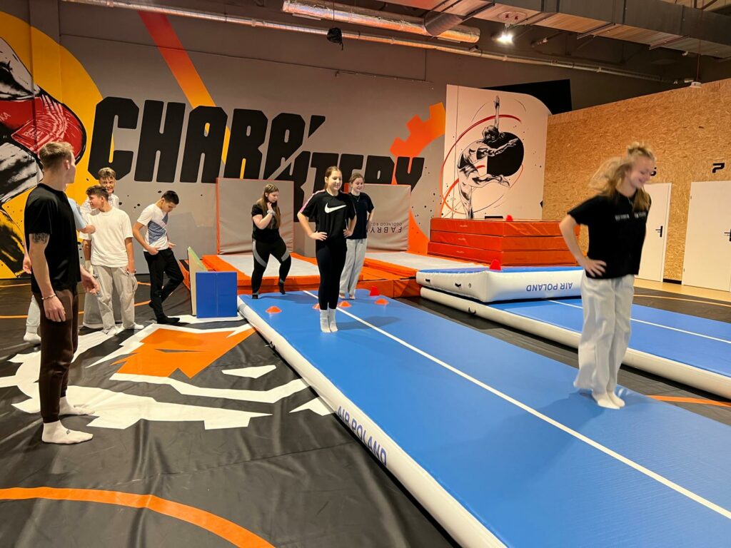 4 dziewczyny wykonujące skoki na ścieżce gimnastycznej obok stoi prowadzący zajęcia oraz 3 chłopaków