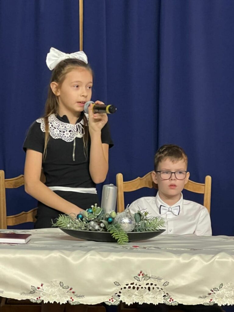 Dziewczynka w czarnej sukience z dużą białą kokardą mówi do mikrofonu przy udekorowanym stole