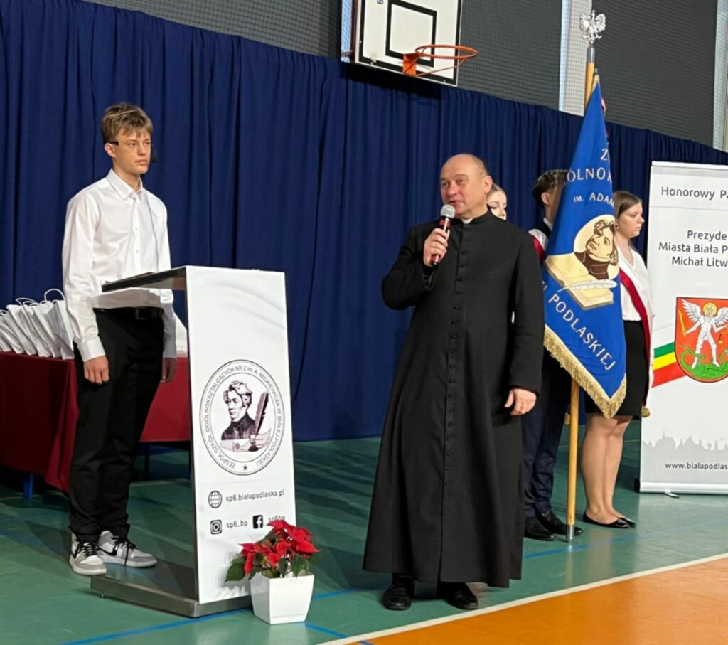 Kapłan przemawia do mikrofonu przy mównicy z logo szkoły, obok stoi uczeń i poczet sztandarowy