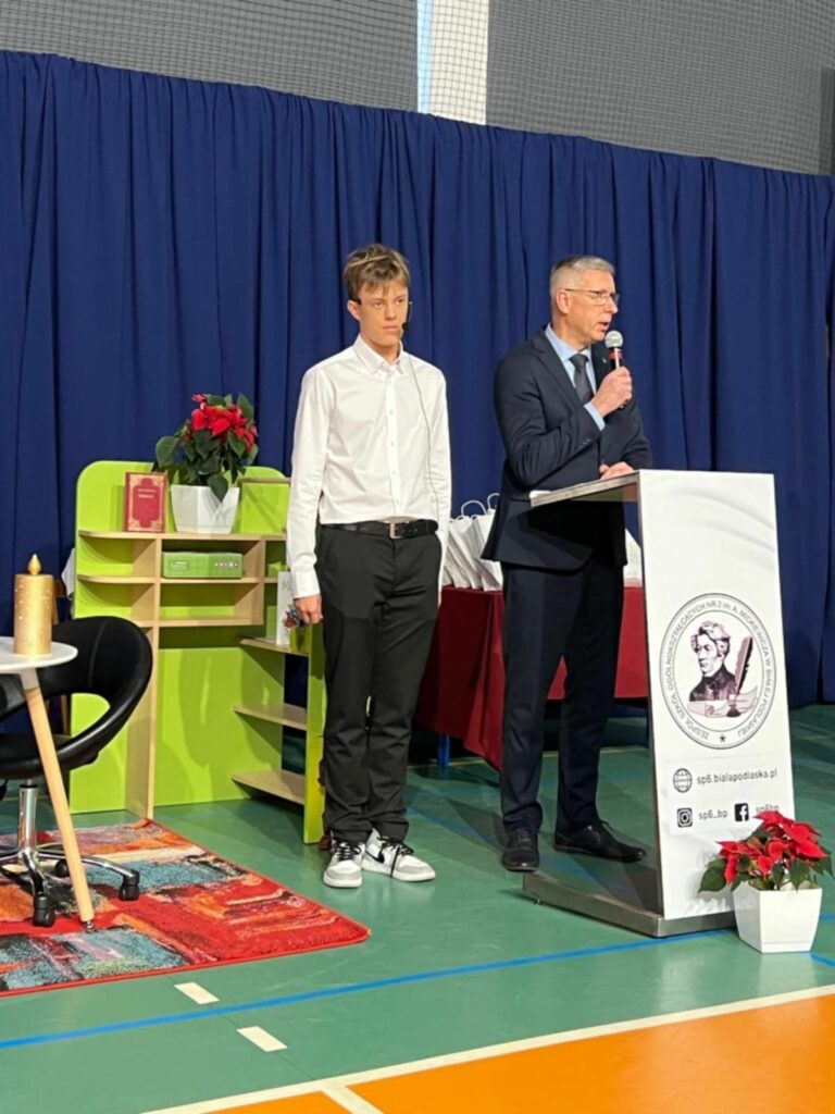 Mężczyzna w garniturze i uczeń stoją przy mównicy, za nimi zielony regał i biurko tworzące elementy scenografii