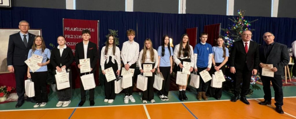 Dyrektorzy i uczniowie stoją w jednym rzędzie na tle choinki i kurtyny, każdy trzyma dyplom i torbę z upominkami