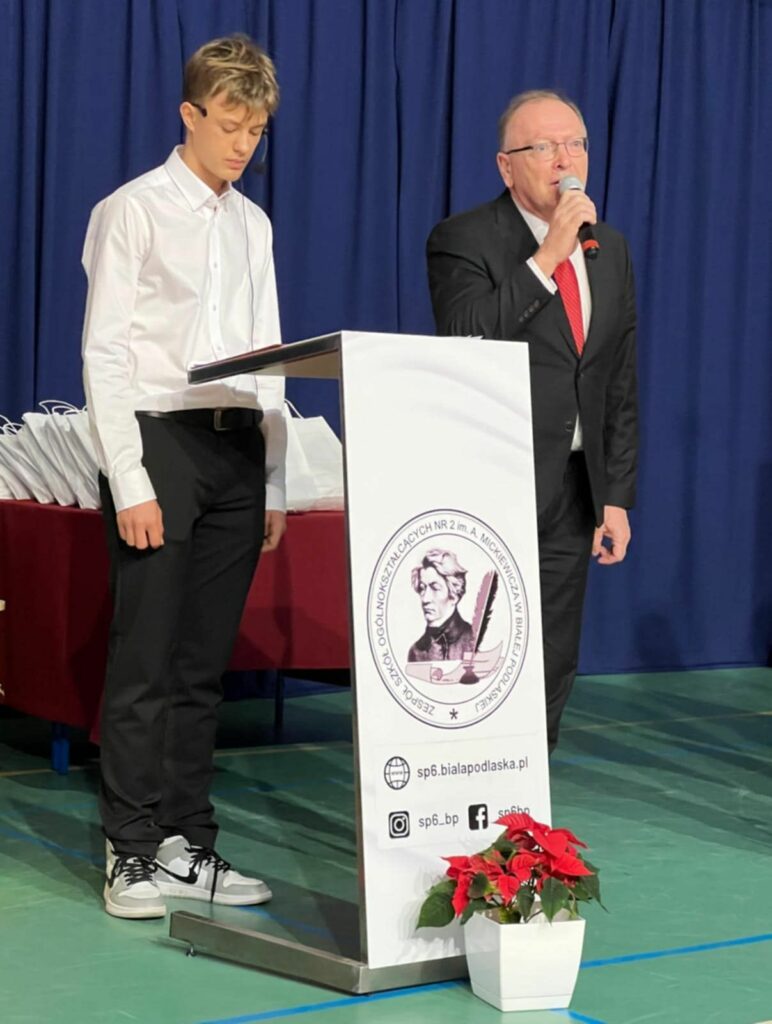 Uczeń i mężczyzna w garniturze stoją przy mównicy z logo szkoły podczas rozdania nagród