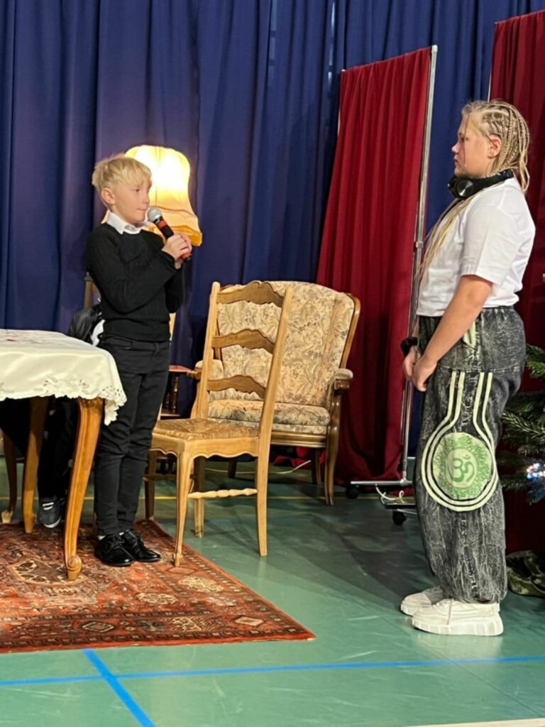 Chłopiec w ciemnym swetrze trzyma mikrofon i zwraca się do dziewczynki w młodzieżowym stroju na tle stołu i fotela