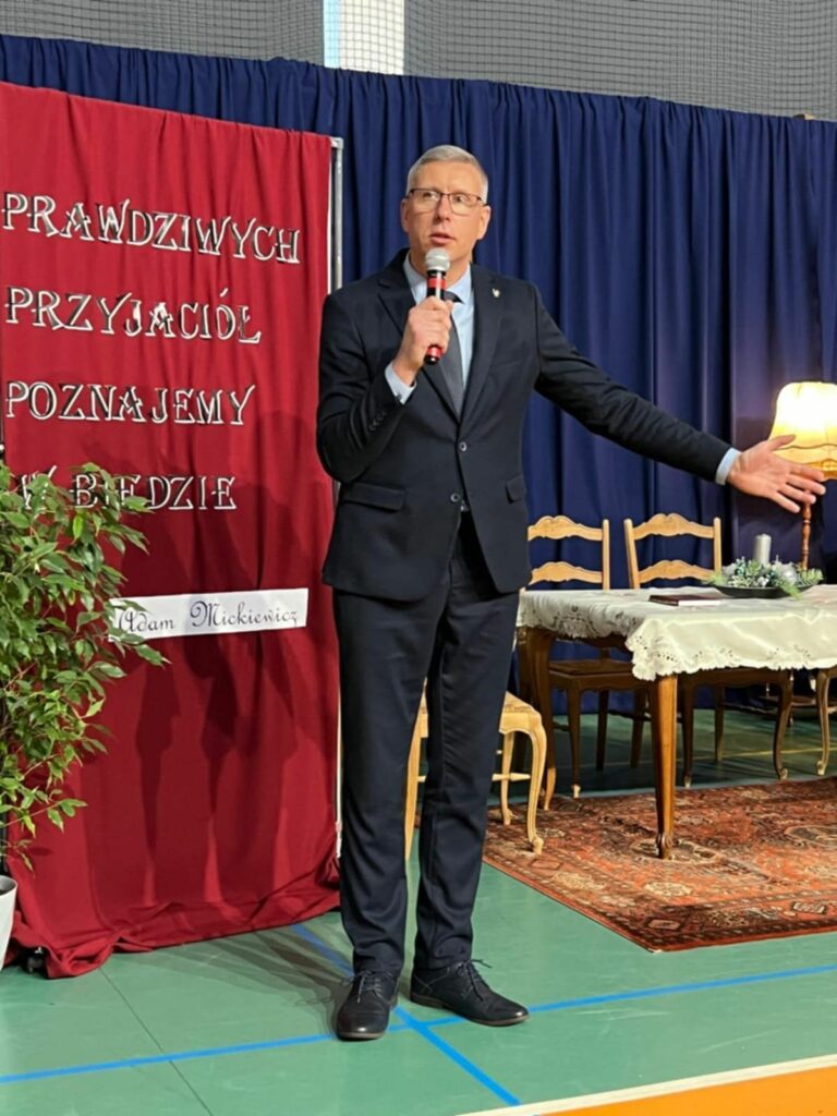 Mężczyzna w ciemnym garniturze przemawia do mikrofonu na tle bordowej planszy z cytatem i szkolnej scenografii