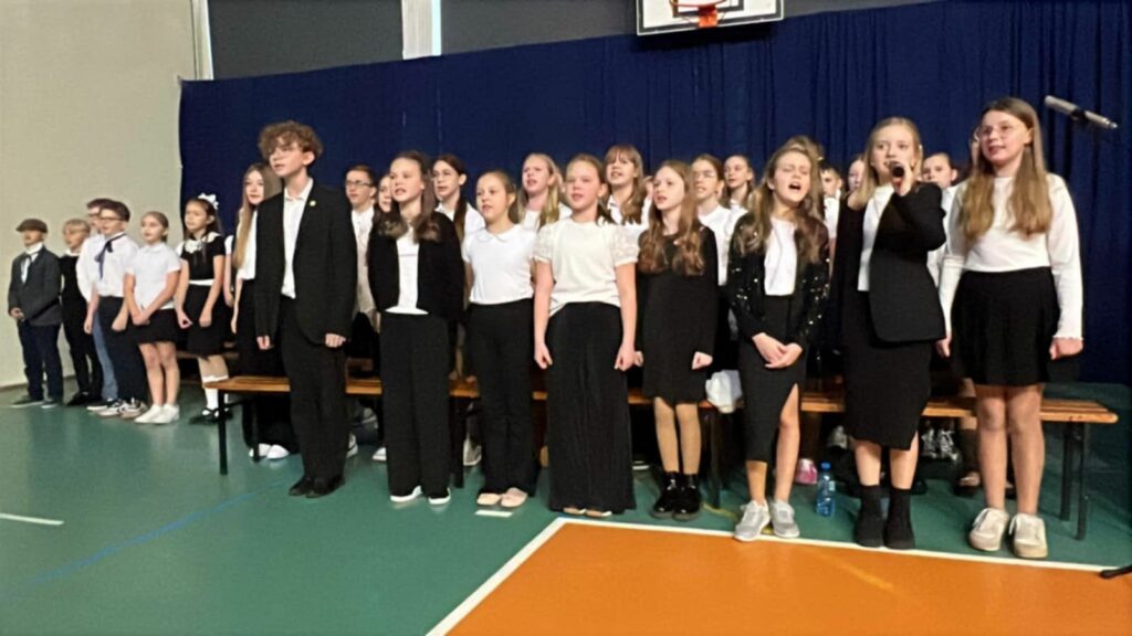 Duża grupa uczniów w białych koszulach i ciemnych spodniach spogląda w stronę aparatu, w tle choinka i scenografia