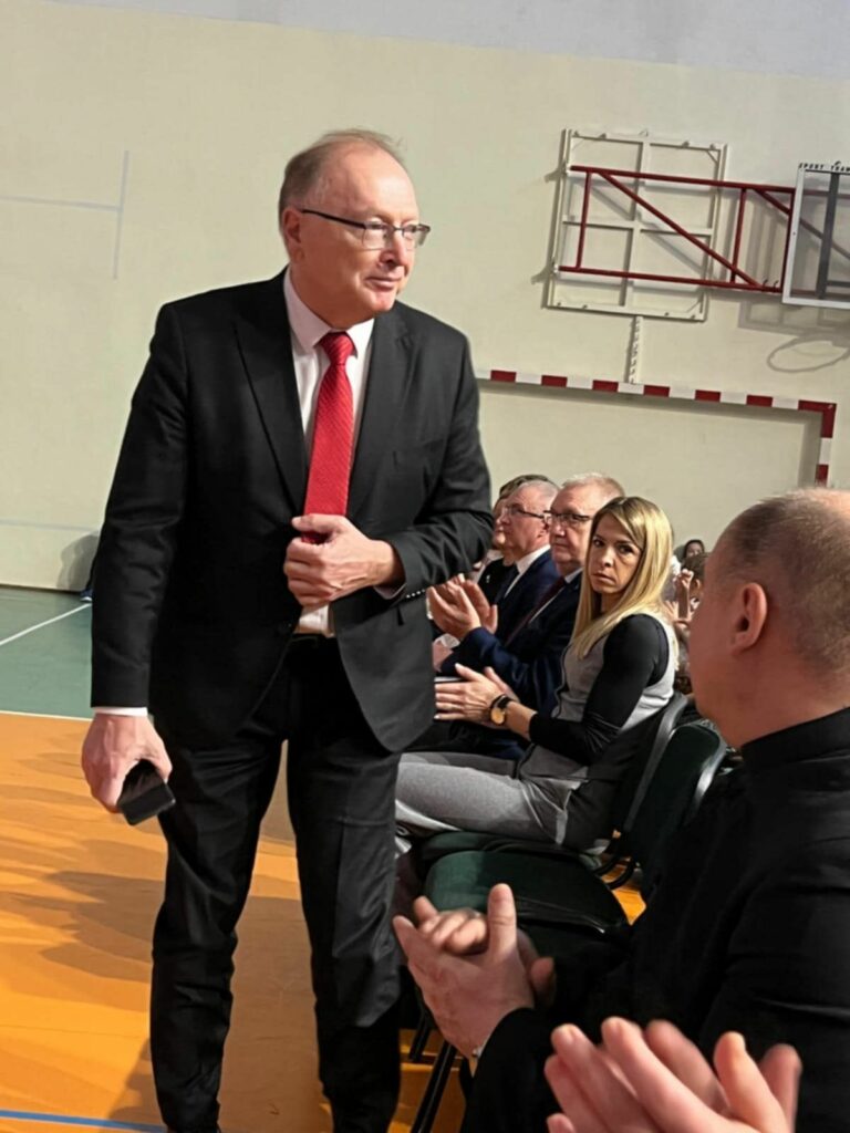 Mężczyzna w garniturze z czerwonym krawacie stoi przed rzędami krzeseł, uczestnicy uroczystości biją brawo