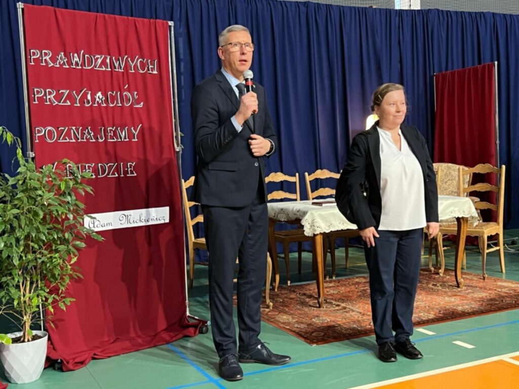 Mężczyzna w garniturze i kobieta w ciemnej marynarce stoją na scenie obok planszy z cytatem Adama Mickiewicza