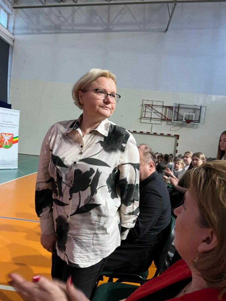 Kobieta w eleganckiej koszuli w kwiaty stoi na sali gimnastycznej podczas szkolnej uroczystości, w tle widownia