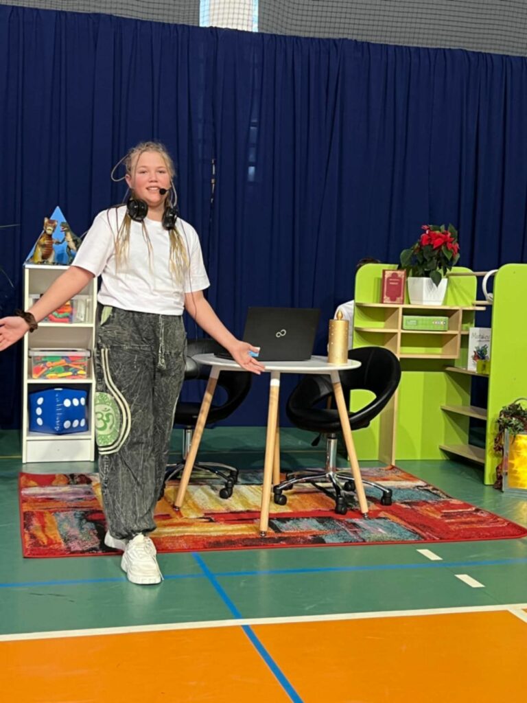 Uczennica w luźnym stroju młodzieżowym stoi na kolorowym dywanie przy biurku z laptopem na tle niebieskiej kurtyny