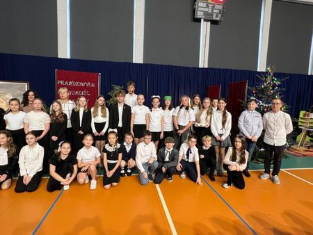 Duża grupa uczniów w białych koszulach i ciemnych spodniach spogląda w stronę aparatu, w tle choinka i scenografia