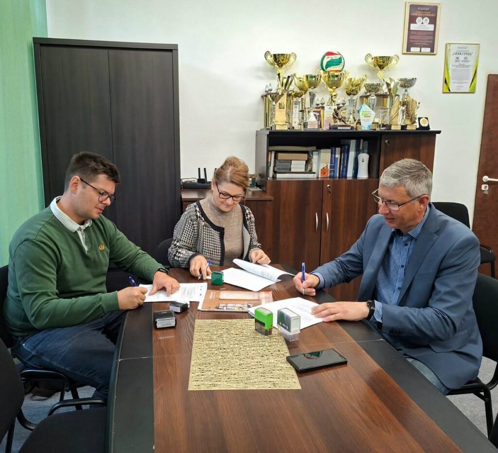 2 mężczyzn i kobieta przy stole podpsują dokumenty na stole leży smartfon i pieczątki