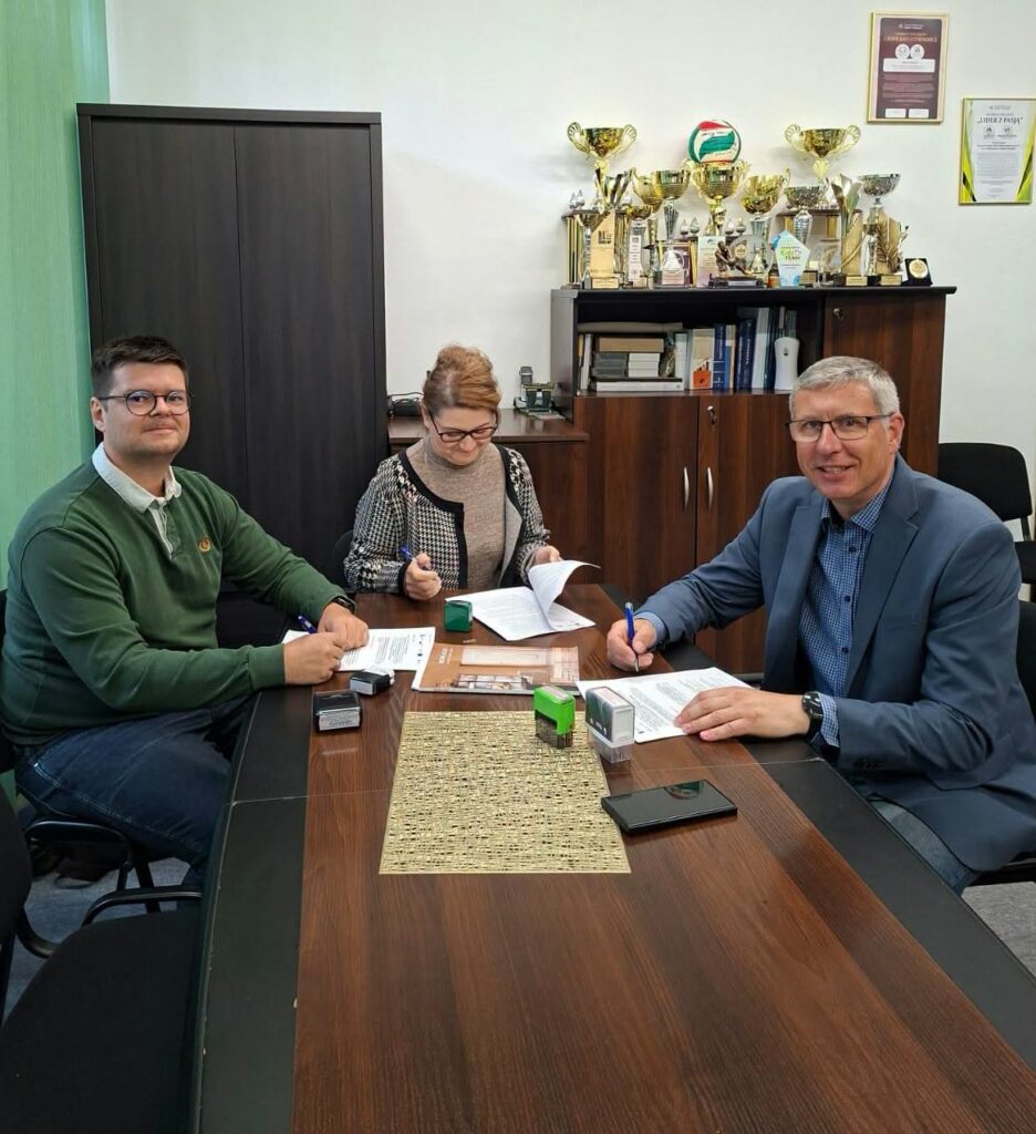 2 mężczyzn podpisując dokumenty patrzy w stronę obiektywu aparatu, kobieta podpsuje dokumenty, na stole leży smartfon i pieczątki